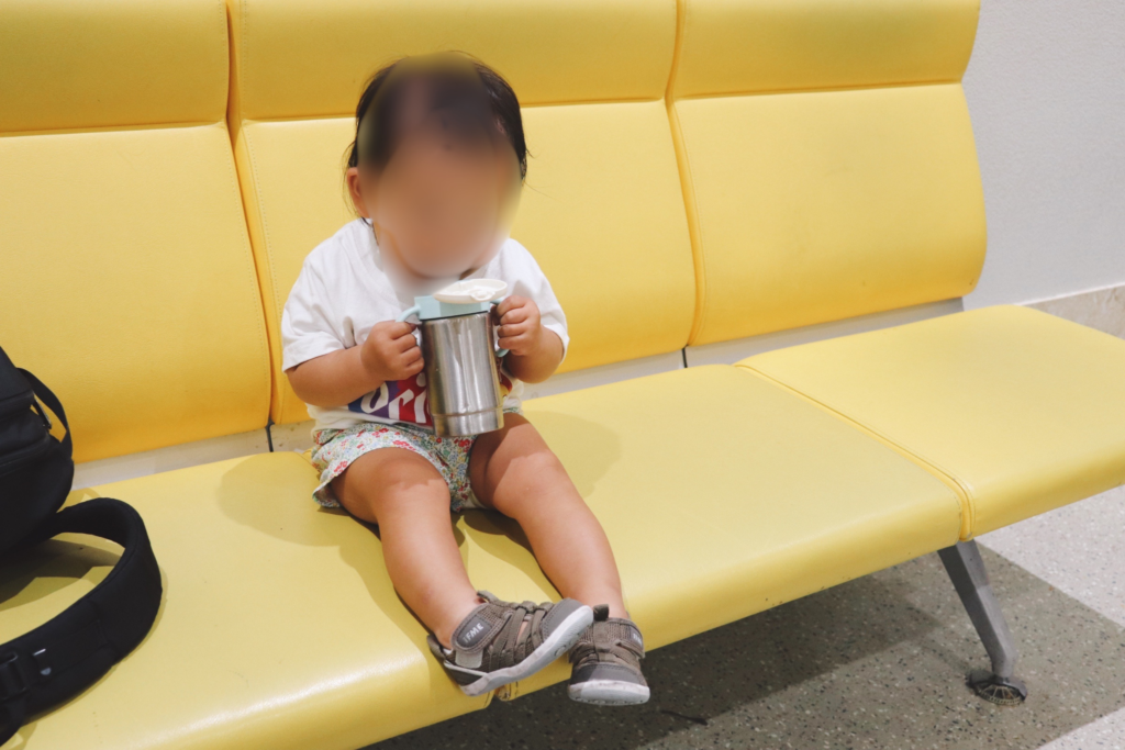 黄色いベンチに座り、両手でふた付きのステンレスマグを持って飲む次女